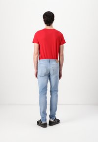 Rode T-shirt met korte mouwen gecombineerd met lichtblauwe jeans. De jeans hebben een beige leren patch op de achterkant en een slim fit ontwerp. Zwarte schoenen.