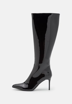 Botte stiletto noire brillante montant jusqu'au genou, avec un bout pointu et un talon haut fin, sur un fond clair uni.
