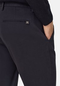 Jūras spēku krāsas kokvilnas bikses ar slaidu piegriezumu, redzamu aizmugurējo kabatu un metāla logo akcentu netālu no kabatas atvēruma.