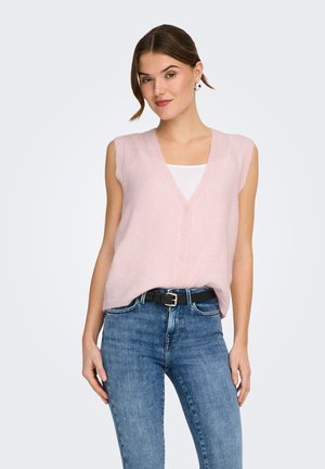 Donna che indossa un gilet senza maniche color rosa chiaro con bottoni sopra una maglietta bianca, jeans blu e cintura nera, in piedi davanti a uno sfondo semplice.