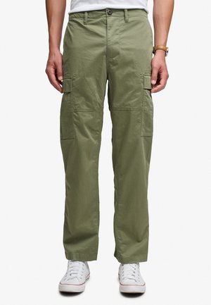 Mann trägt olivgrüne Cargohose mit Klappentaschen und weißen Sneakers, steht vor einem schlichten weißen Hintergrund.