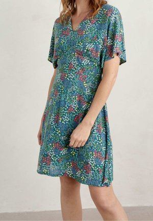 Žena v krátkých rukávech, zelených šatech délky ke kolenům s barevným květinovým potiskem stojí na jednoduchém světlém pozadí.