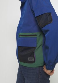 Färgglad vindjacka med en blå ovandel, grön nederdel och mesh-accenter, samt flera dragkedjefickor och patchfickor.