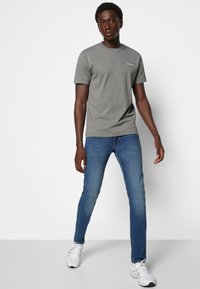 Camiseta gris de manga corta con cuello redondo y un pequeño logo en el pecho, combinada con jeans de mezclilla azules y zapatillas blancas.