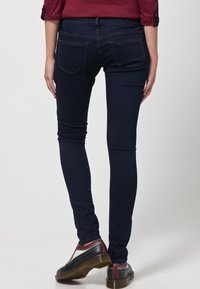 Vaqueros ajustados de mezclilla azul oscura, con una cintura de media altura, bolsillos traseros y una textura suave. Combinados con zapatos marrones con detalles.