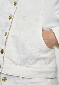 Vit denimjacka med präglade mönster av hjärtan och initialer, guldfärgade knappar och en figursydd design med en lätt texturerad yta.