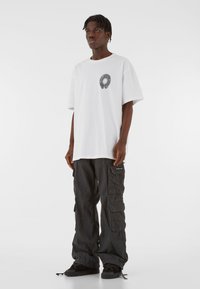 Biele oversize tričko s kruhovou čiernou grafikou na hrudi, spárované s čiernymi cargo nohavicami s viacerými vreckami a značkovými akcentmi.