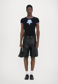 Schwarzes, figurbetontes T-Shirt mit lila Akzenten und Sternenmotiv, kombiniert mit schwarzen Leder-Shorts mit Reißverschlüssen und einer strukturierten Handtasche.