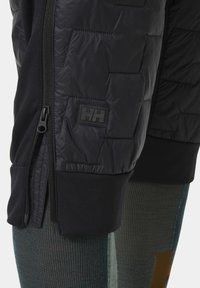 Czarne szorty izolowane o pikowanej strukturze, z zamkniętą kieszenią po boku i naszywką z logo. Pod spodem widoczne są zielone legginsy o teksturowanej powierzchni.