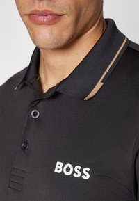 Camiseta polo negra con cuello acanalado y acentos beige, dos botones y logo blanco "BOSS" impreso en el pecho. Tejido suave y texturizado.