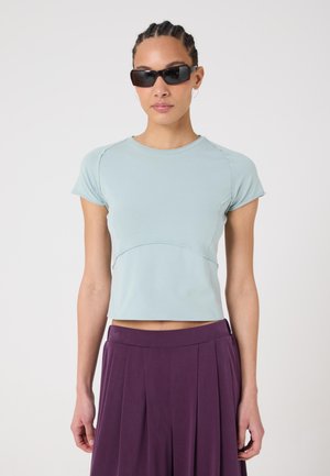 Frau trägt hellblaues, figurbetontes Kurzarmshirt, dunkelvioletten, plissierten Rock und schwarze, rechteckige Sonnenbrille, steht vor weißem Hintergrund.