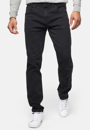 Homme portant un pantalon noir slim et des baskets blanches, marchant vers l'avant sur un fond clair uni.