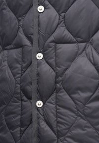 Καπιτονέ μαύρο γιλέκο με απαλή υφή, τέσσερις κουμπότρυπες και δύο μπροστινές τσέπες, διακοσμημένο με ορατά μοτίβα ραφής.