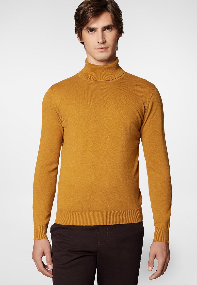 Musztardowy sweter z golfem wykonany z miękkiej, prążkowanej tkaniny, z długimi rękawami i dopasowanymi mankietami, w połączeniu z ciemnobrązowymi spodniami.