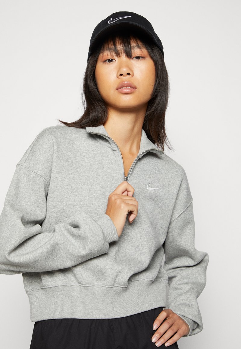 Sudadera gris recortada con cuello con cremallera y cinturilla elástica; cuenta con un logo blanco de Nike en el pecho. Gorro negro con un logo blanco.