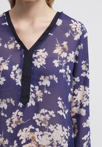 Blouse en voile marine avec un motif floral en crème et beige, dotée d'un décolleté en V et d'un ourlet noir. Tissu léger à la texture fluide.