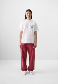 T-shirt bianco in cotone con testo grafico verde sul petto, abbinato a pantaloni larghi rosso-rosa e sneaker bianche.