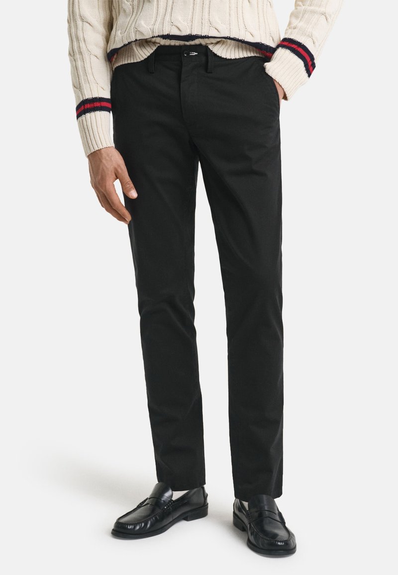 Schwarze Chinos mit geradem Schnitt, die eine glatte Textur und Standardtaschen aufweisen. Kombiniert mit einem cremefarbenen gerippten Pullover und schwarzen Slippern.