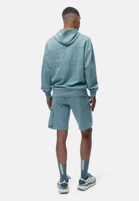 Hele sinine ökoloogilise disaini kapuutsiga sviiter, sobivad lühikesed püksid ja mustrilised sokid. Omab vabalt voolavat siluetti ja tekstuurses detailides kangas.