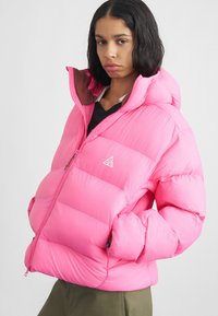 Chaqueta rosa acolchada con capucha, presenta un logo en el pecho, bolsillos con cremallera y una tela texturizada. Diseño suave y aislante.