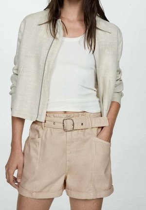 Femme portant un short beige ceinturé, un débardeur blanc côtelé et une veste courte beige clair zippée, les mains dans les poches.