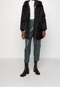 Parka imbottita nera con cappuccio staccabile, dotata di cerniera frontale, tasche laterali e polsini a costine. Indossata sopra pantaloni verdi in ecopelle e stivali neri.