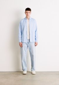 Lichtblauw overhemd met knopen over een crèmekleurig T-shirt, gecombineerd met lichtblauwe jeans en witte sneakers. Eenvoudig, casual ontwerp met een royale pasvorm.