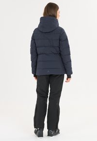 Marineblaue gepolsterte Jacke mit Kapuze, vertikaler Steppung und elastischen Bündchen, kombiniert mit schwarzen Schneehosen. Klarer, wetterfester Entwurf.