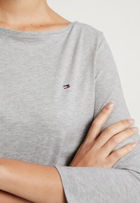 Graues Langarmshirt mit Bootsausschnitt, weichem Material und einem kleinen gestickten Logo in Rot, Weiß und Blau auf der Brust.