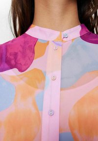 Blusa colorata con forme astratte in viola, arancio e blu, dotata di collo rialzato e chiusura frontale con bottoni nei colori abbinati.
