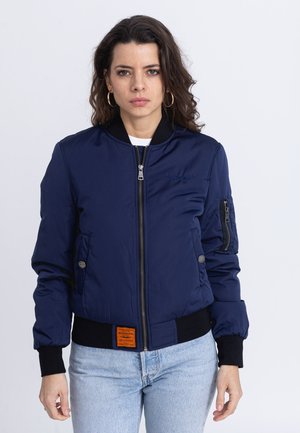 Woman with curly dark hair wears a navy blue bomber jacket with black cuffs and light blue jeans, standing against a plain white background.