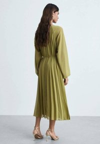 Rochie plisată de culoare verde măslinie cu mâneci lungi, talie strânsă și material fluid. Sandale aurii cu curele completează ținuta.