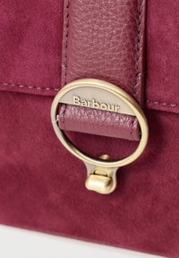 Bordó velúr kézitáska bőr pánttal, sárgaréz hardware-rel és "Barbour" felirattal gravírozott körgyűrűvel. Texturált felület és varrási részletek.