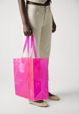Transparente pinke Tragetasche mit kontrastierenden neonpinken Trägern, rechteckiger Form und glänzender Oberfläche, gehalten von einer Person in hellen Hosen.