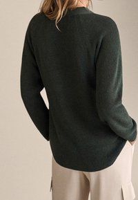 Personne portant un pull en tricot côtelé vert foncé à manches longues et un pantalon beige, vu de dos.