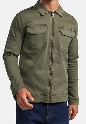 Mand iført olivengrøn lynlåstrøje med to klaplommer på brystet og blå jeans, vist fra nakke til hofter.
