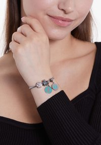Zilveren bedelarmband met een bloemaccent, turquoise schijven met figuren en een donkere edelsteen. Draag het om een pols met een gestructureerde zwarte top.