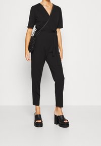Mujer vestida con un mono negro con envoltura, sandalias de plataforma con tacones, bolso de malla cruzado y collares de plata en capas, de pie contra un fondo blanco.