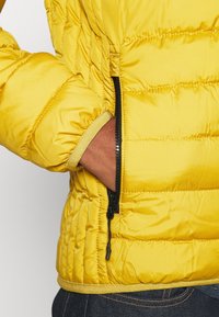 Main dans la poche d'une veste matelassée jaune vif avec une fermeture éclair noire, portée sur un jean denim foncé.