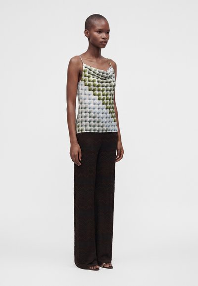 Femme mince aux cheveux courts portant un haut sans manches à motifs verts et blancs et un pantalon noir à jambes larges, debout devant un fond uni.