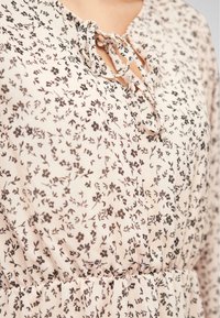 Robe fleurie de couleur rose clair avec un motif floral noir. Elle possède une fermeture à nouer au niveau du col et une taille froncée. Tissu à la texture douce.