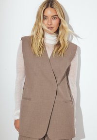 Rjava bezrokavna jopica z oblikovanim krojem, ki vključuje dve sprednji žepi in subtilno teksturo, je složena preko svetlega turtleneck puloverja.