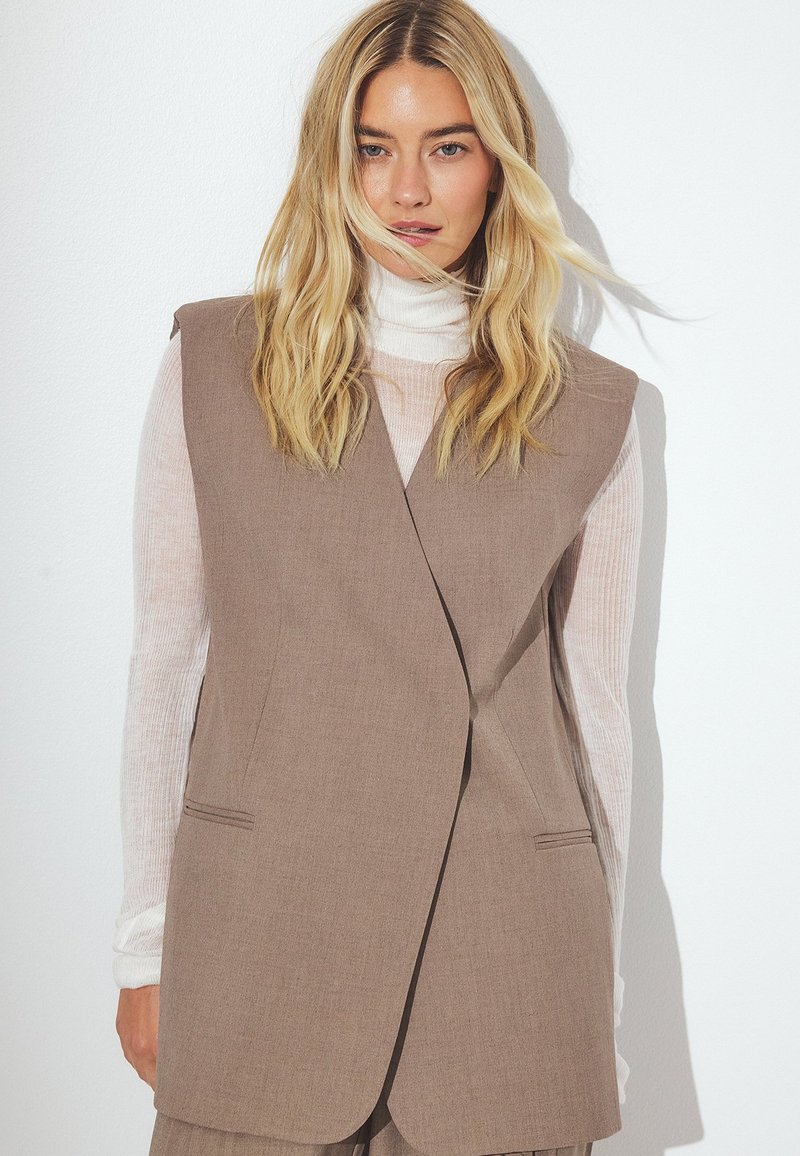 Rjava bezrokavna jopica z oblikovanim krojem, ki vključuje dve sprednji žepi in subtilno teksturo, je složena preko svetlega turtleneck puloverja.