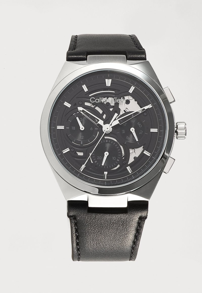 Ρολόι χειρός Calvin Klein με μαύρο δερμάτινο λουράκι, ασημί κάσα και μαύρο σκελετό καντράν που δείχνει ώρα, ημερομηνία και υποκαντράν ημέρας.