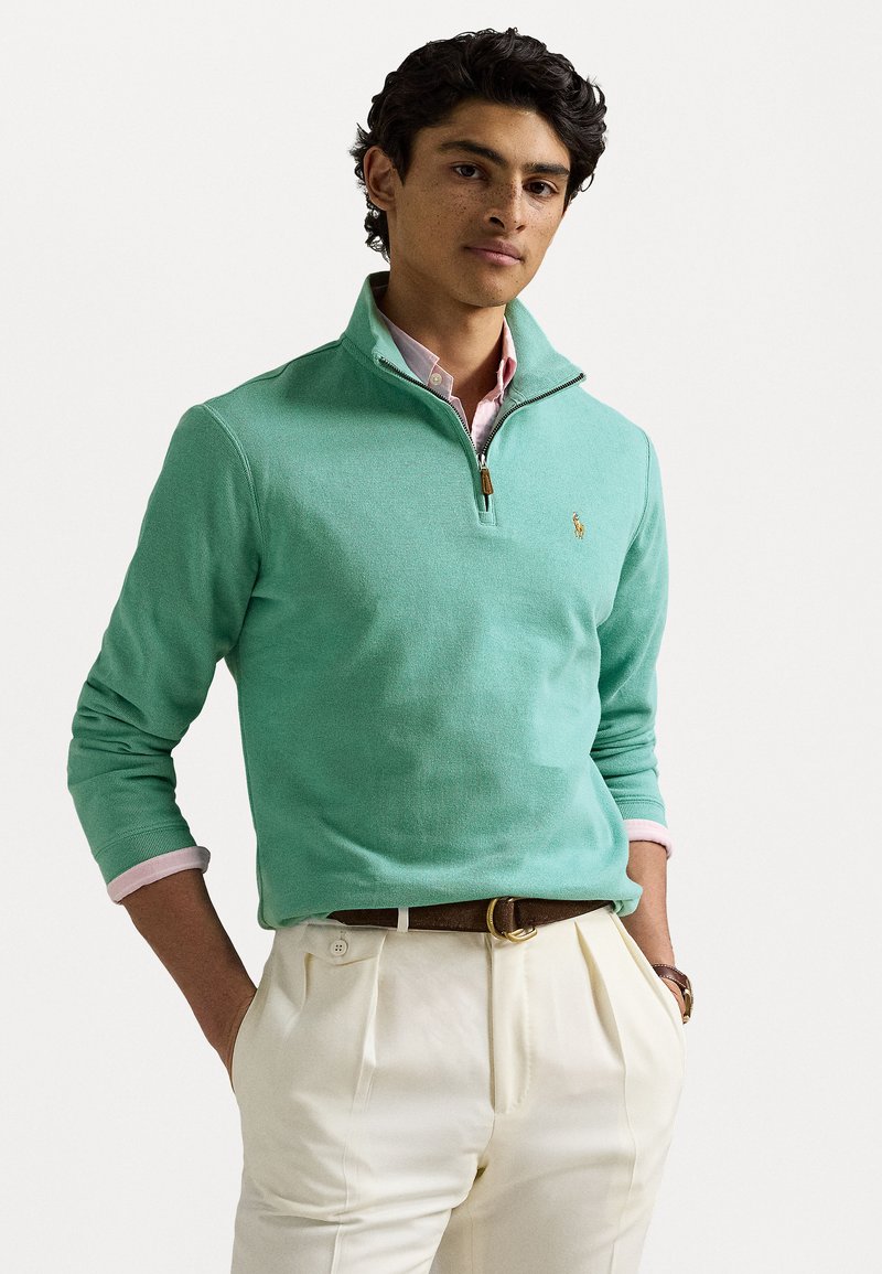 Πράσινο μέντας quarter-zip pullover από υφασμα με υφή. Διαθέτει χρυσό φερμουάρ και κεντημένο λογότυπο, φορώντας πάνω από ένα ροζ πουκαμίσα με γιακά.