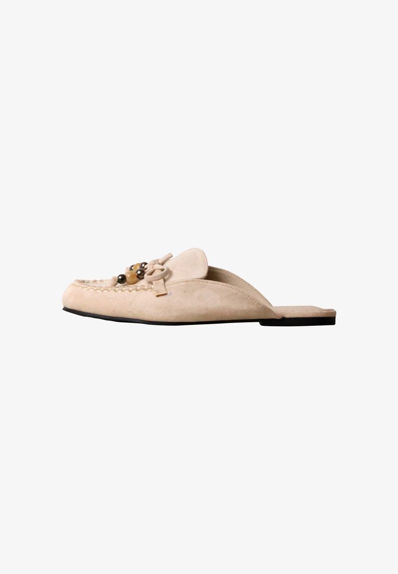 Mule en daim beige à enfiler avec un nœud décoratif et des perles en bois, semelle plate noire et couture style mocassin sur l’orteil.