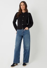 Cardigan nero con scollo rotondo e bottoni frontali, abbinato a jeans larghi blu e sneakers nere. Outfit semplice e casual.