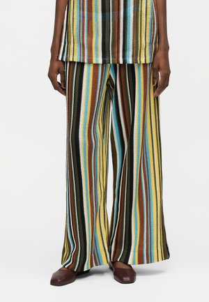 Άτομο που φοράει πολύχρωμο παντελόνι καμπάνα με κάθετες ρίγες και ταιριαστό τοπ, σε συνδυασμό με σκούρα καφέ παπούτσια με τετράγωνη μύτη, στέκεται πάνω σε λευκό φόντο.