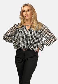 Czarna i biała bluzka w wzór houndstooth z przezroczystymi rękawami, głębokim dekoltem w literę V oraz złotym akcentem w postaci łańcuszka, zestawiona z czarnymi spodniami.