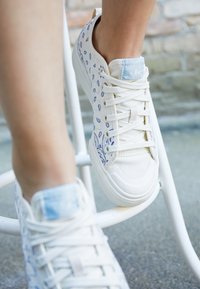 Vita canvas-sneakers med blå paisleymönster, rund tå och platt vit gummisula. Snörningarna är vita, med en ljusblå accent på tungan.